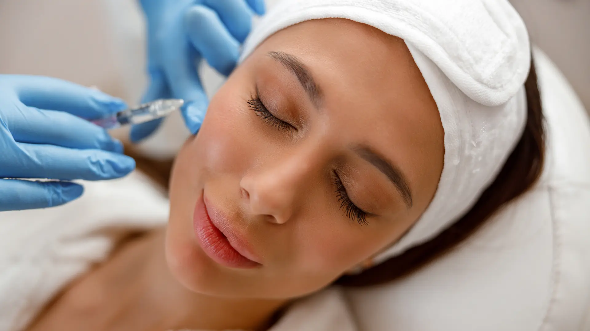 A woman wearing a white headband lies with her eyes closed while a gloved hand administers an injection to her face. The setting appears to be a clinical or spa environment, and the woman looks relaxed.