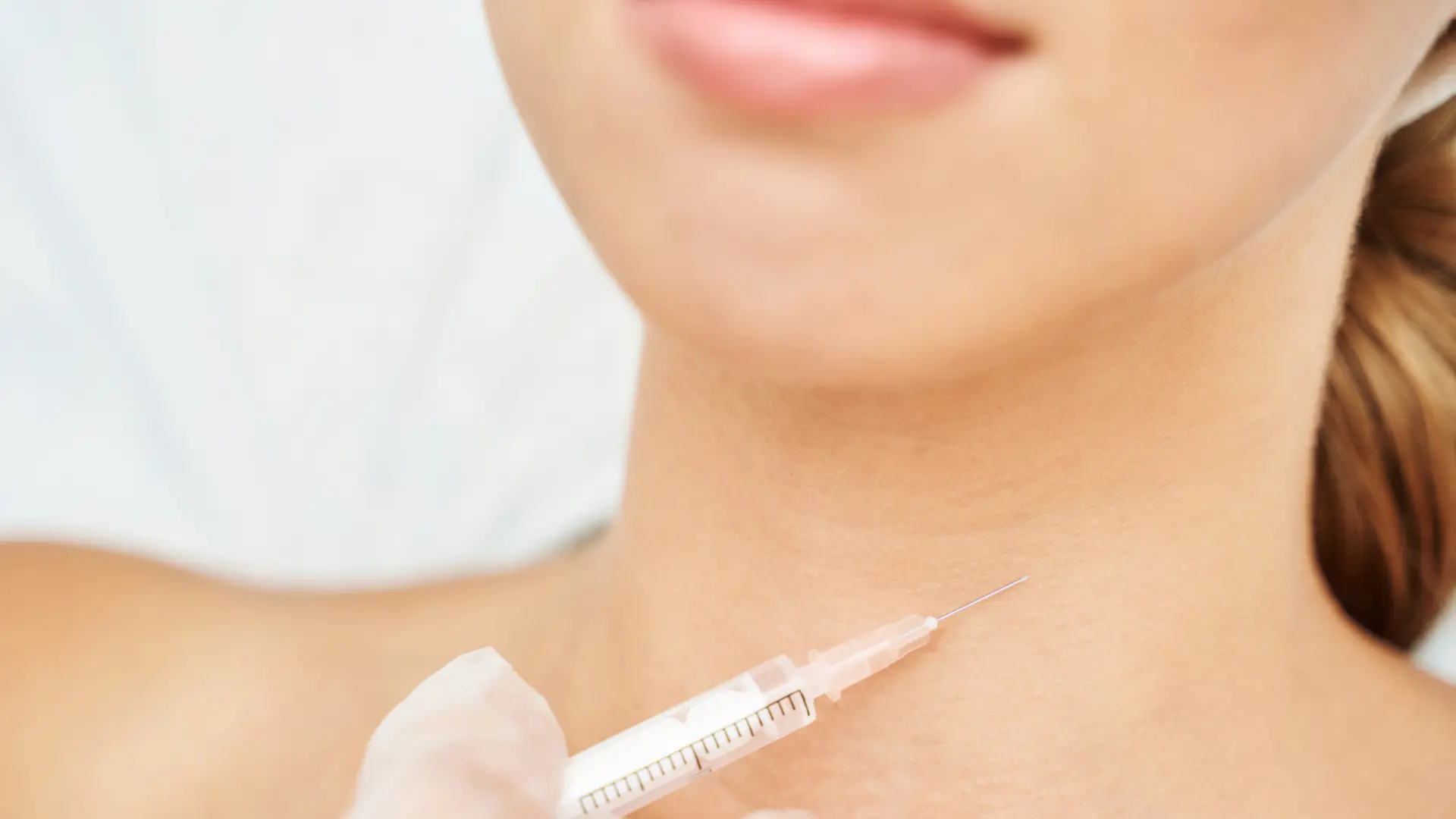 Close-up of a person receiving an injection in the neck area. The image captures gloved hands holding a syringe with a fine needle, while the person's neck and part of the chin are visible. The background is out of focus, emphasizing the injection procedure.